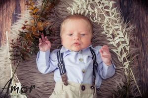 Baby wearing suspenders and bow tie lying on fluffy rug with autumn decorations.