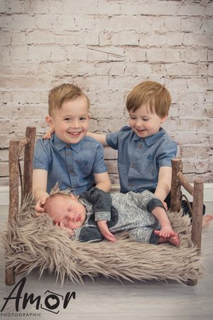 Three siblings sitting on wooden bench against rustic brick wall backdrop.