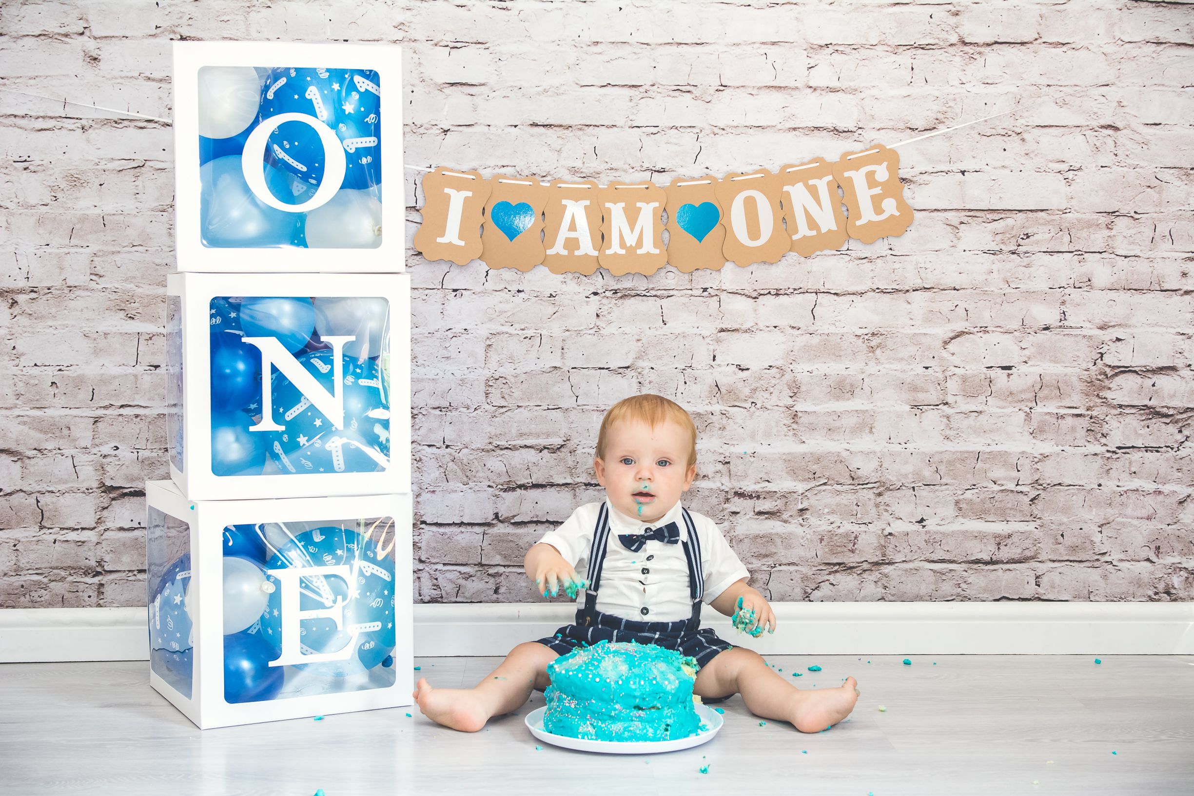 Baby in blue celebrating first birthday with cake and decorative blocks against brick wall.