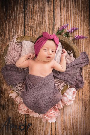 Baby wearing pink headband lying on wooden backdrop with purple flowers and soft textiles.