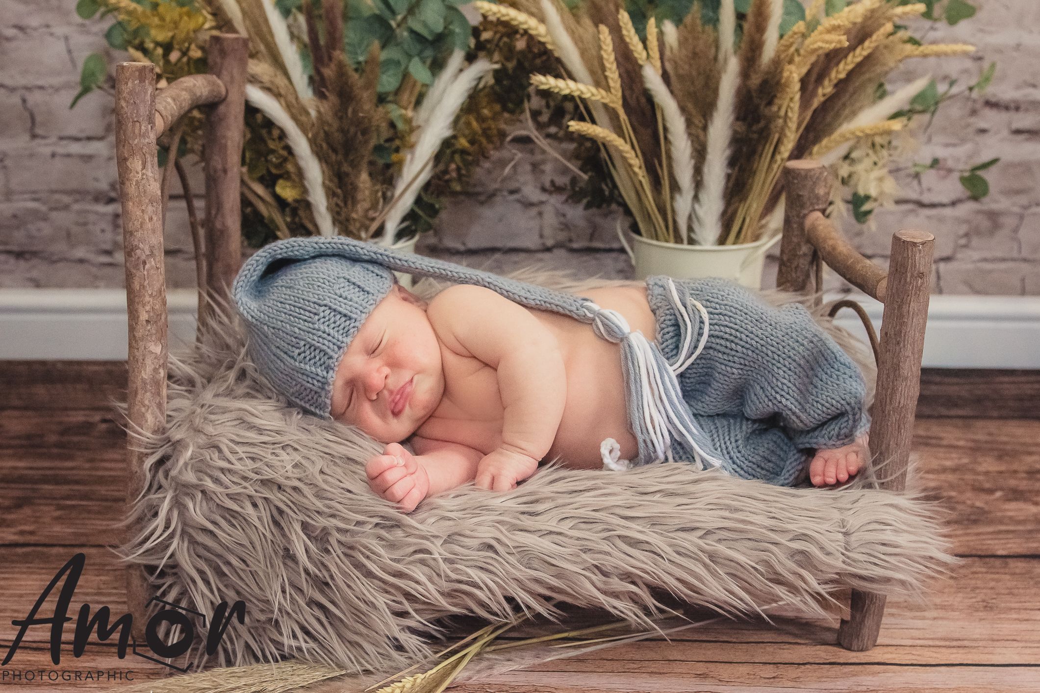Artistic newborn portrait on furry grey blanket with dried wheat stalks and brick background.