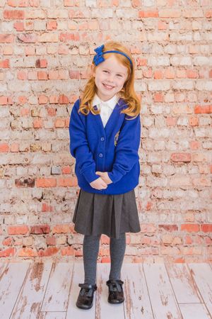 Student in blue cardigan and grey uniform poses against brick wall backdrop.