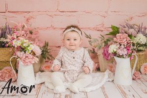 Baby in white outfit sitting among vintage floral arrangements against pink brick wall.