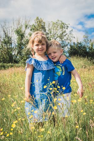 Two children in blue outfits standing together in wildflower meadow on sunny day.