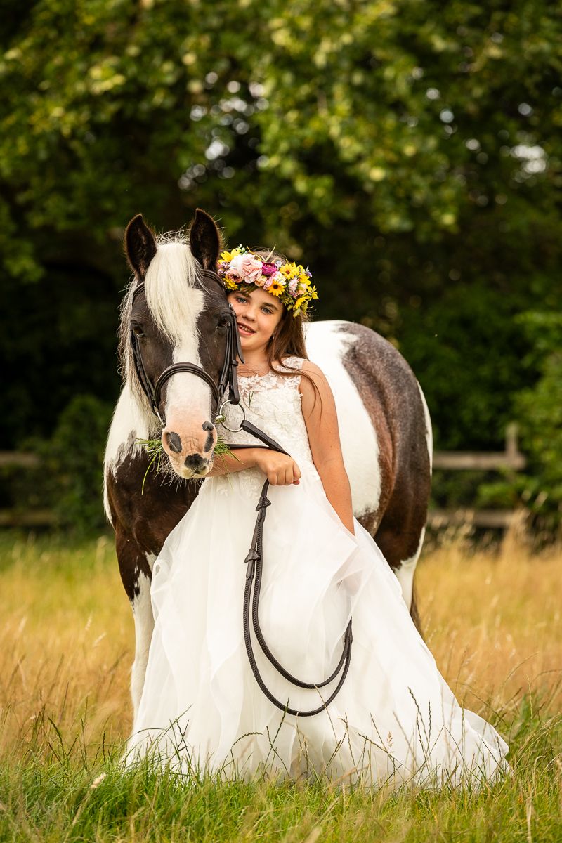 A black and white horse stands with someone in a white dress and floral crown in a summer meadow.