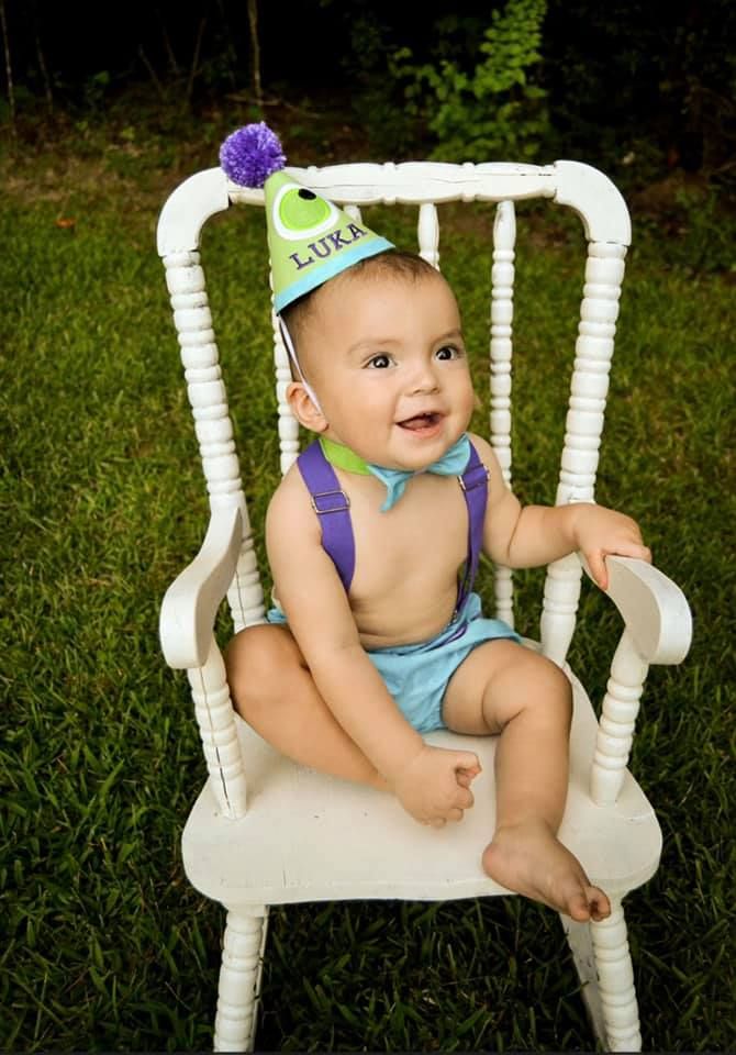 Baby sits in white wooden chair wearing blue outfit and purple accessories.