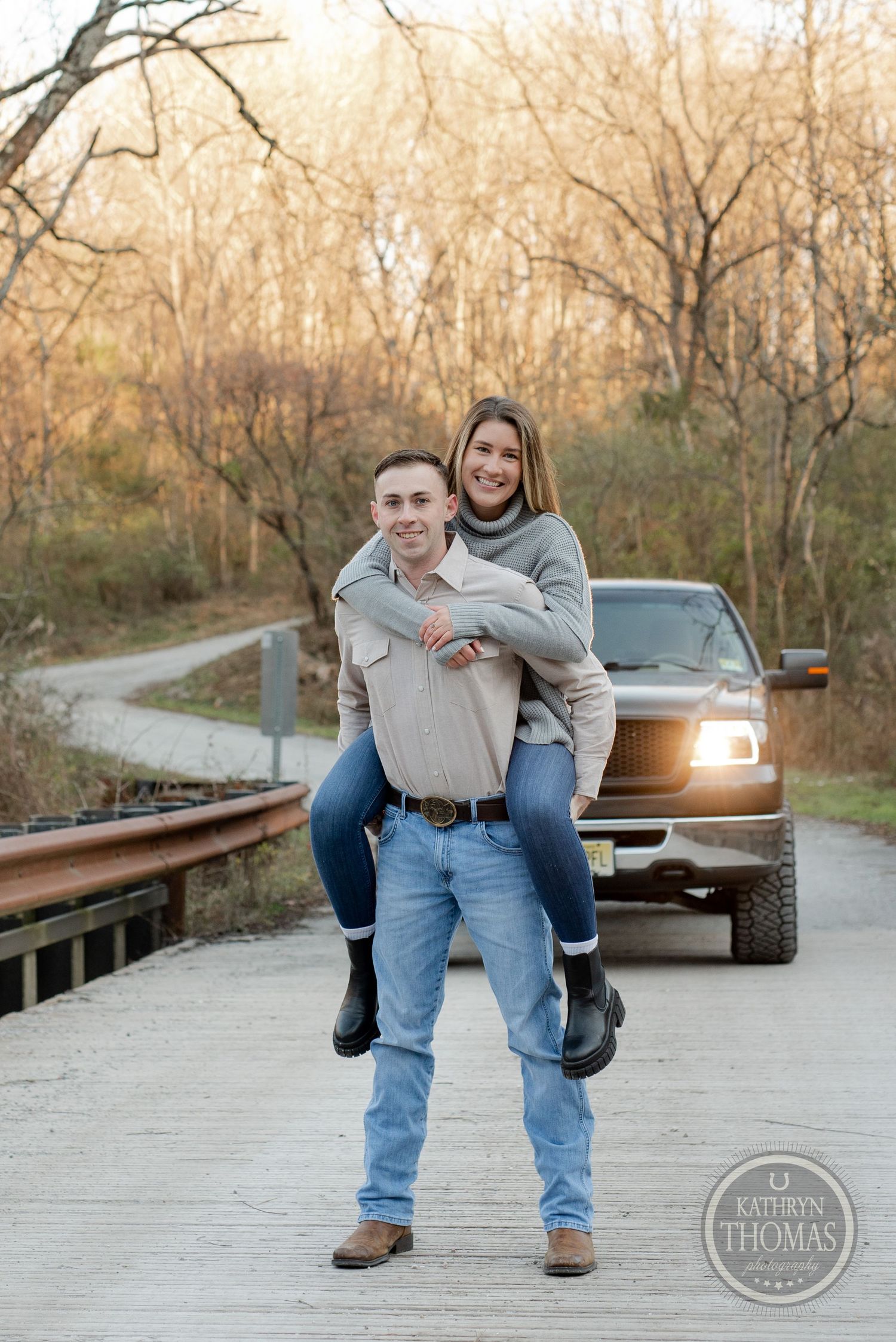 Playful piggyback pose on wooden bridge with pickup truck and autumn trees in background.