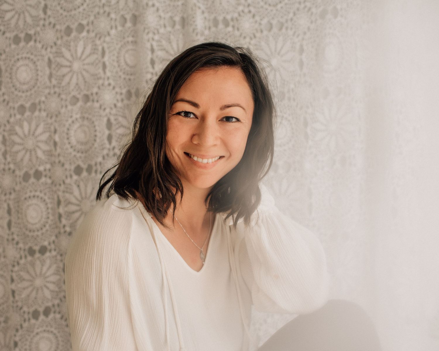 A woman in a white blouse smiling warmly against a textured wall background.