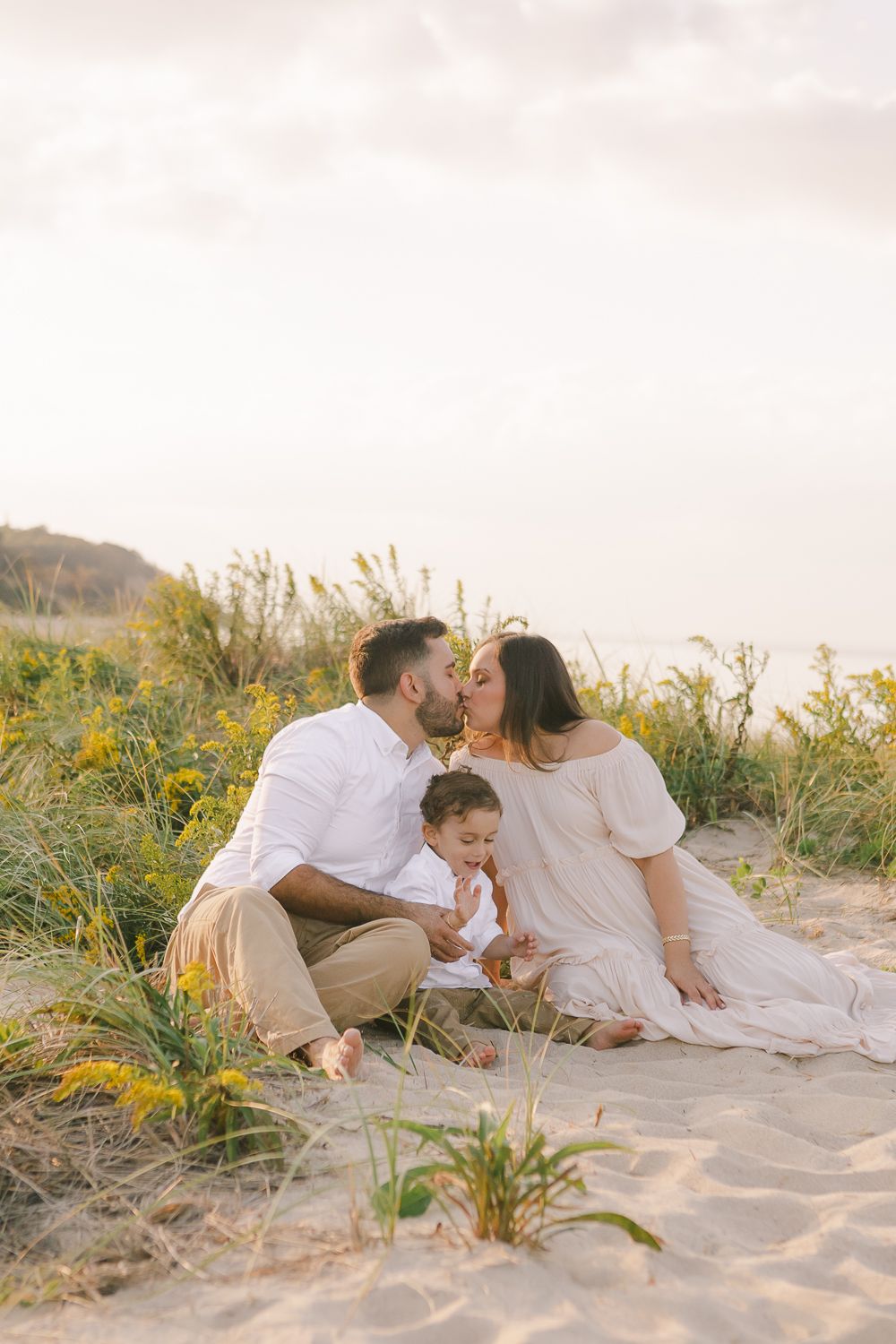 a candid view of a couple sitting on the sand with grass on the background and beautiful light while sharing a kiss