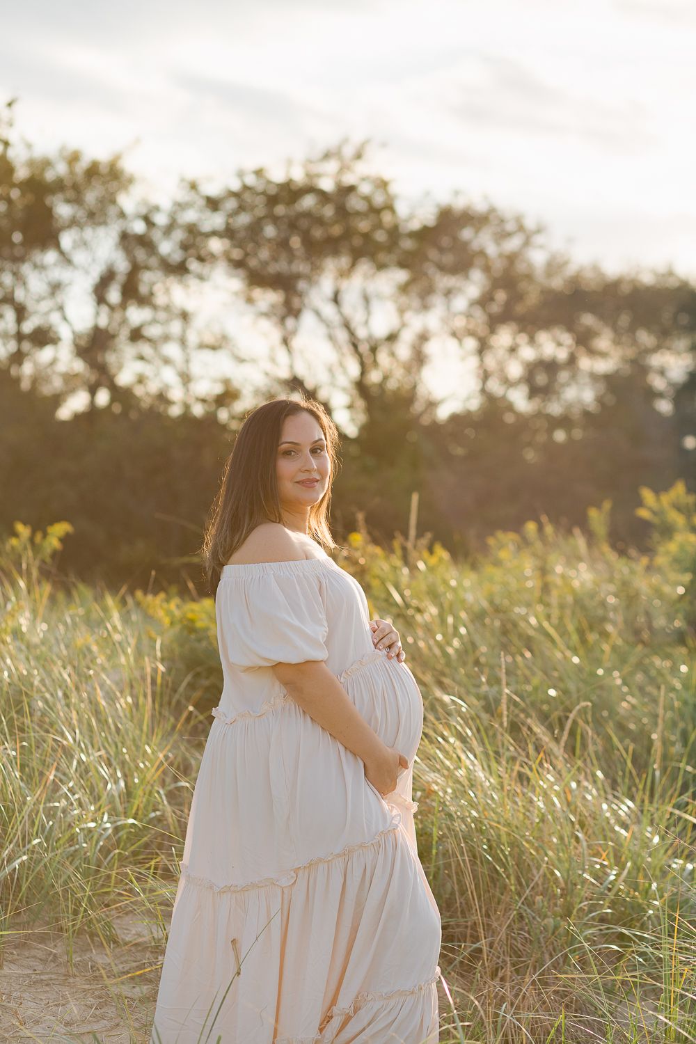 a profile view of a pregnant woman during golden hour wearing a beige dress against a grass background at the beach