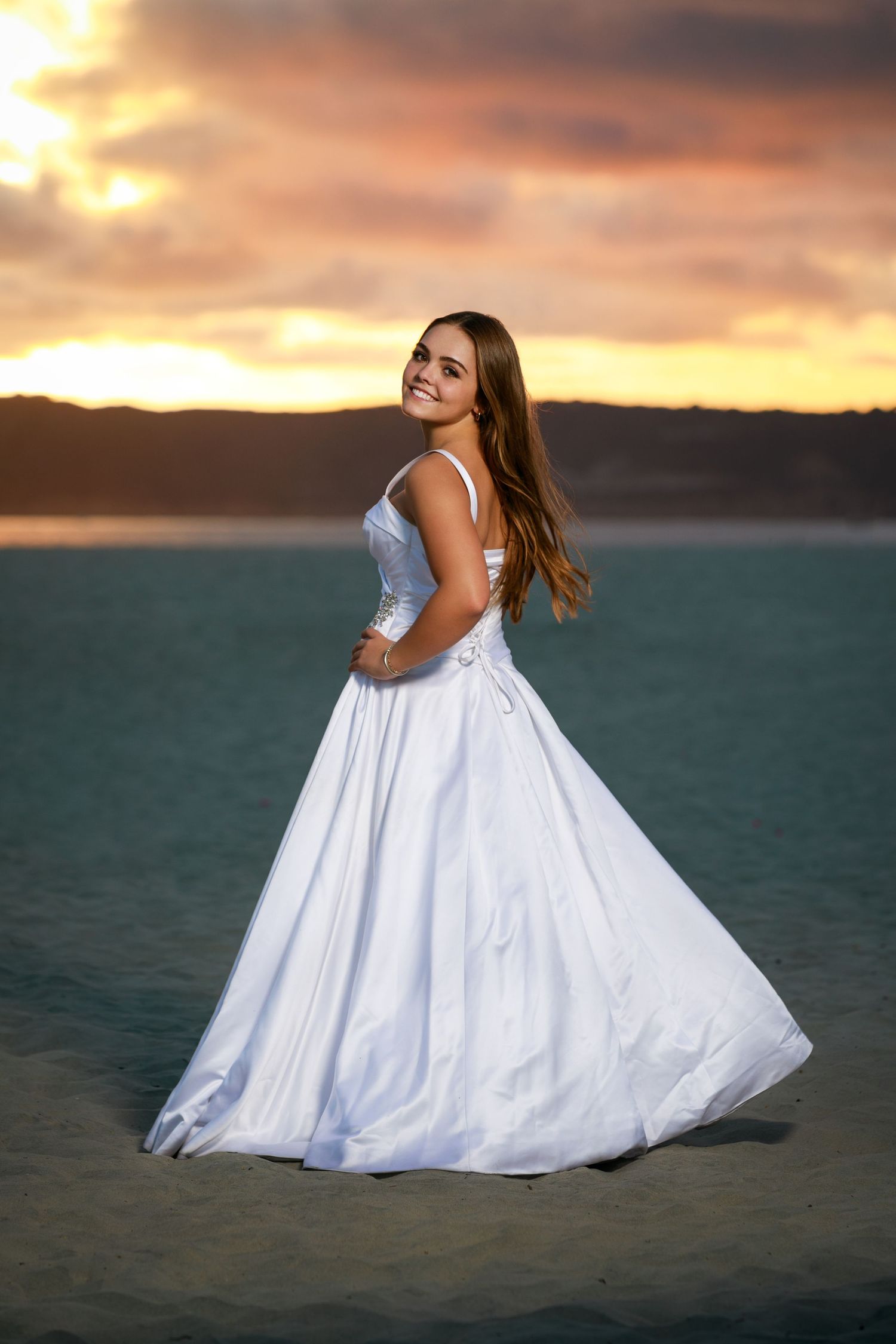 A person in a white gown twirls against a dramatic sunset sky over water.