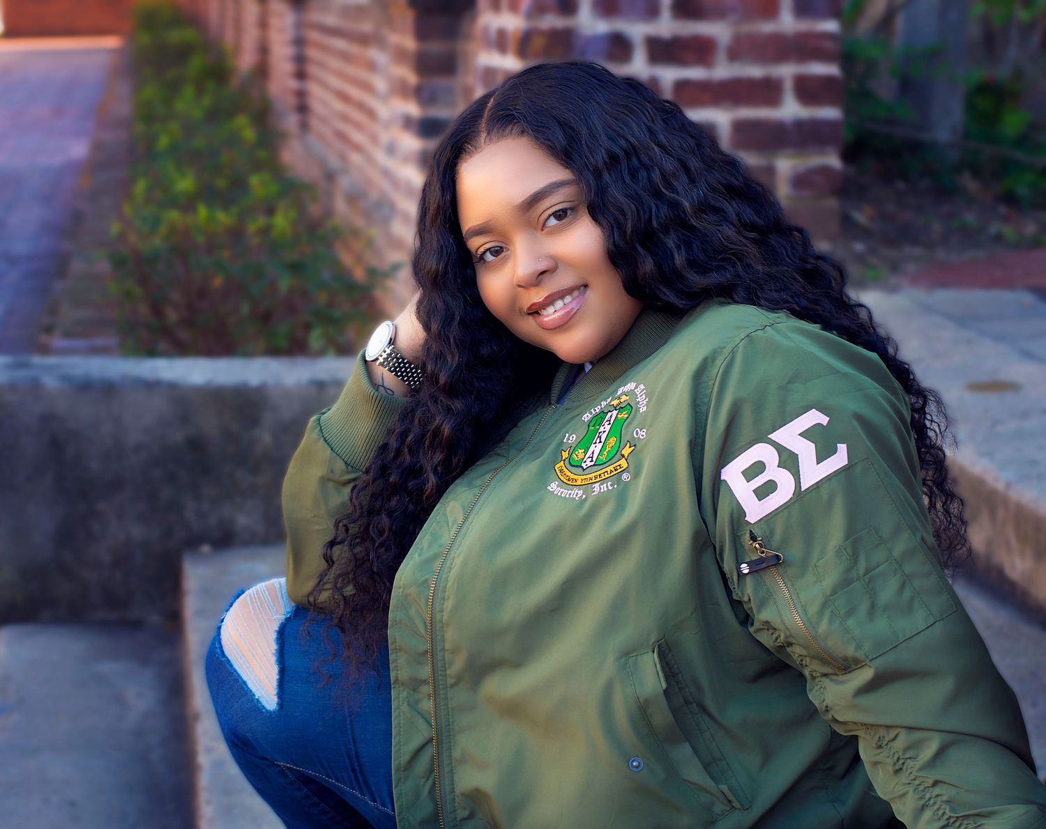 Casual portrait in green varsity jacket against brick wall background.