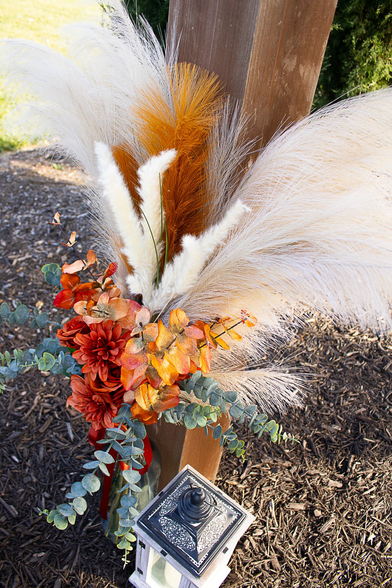 Fall-themed floral arrangement with orange lilies and white feathers against wooden post.