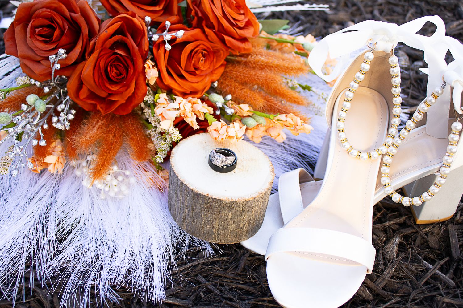 Wedding details with orange roses, white feathers, and pearl-adorned bridal shoes.