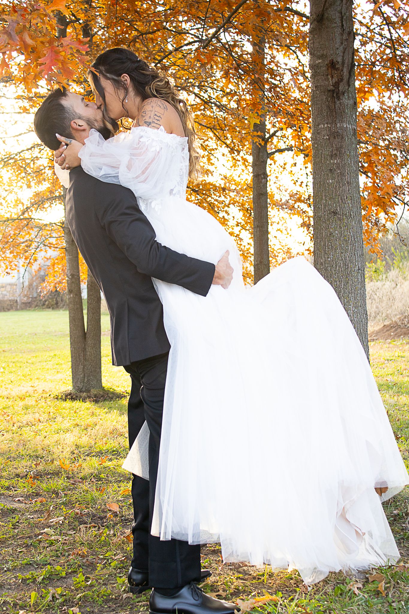 A romantic wedding photo of a couple kissing under autumn trees with golden leaves at sunset.