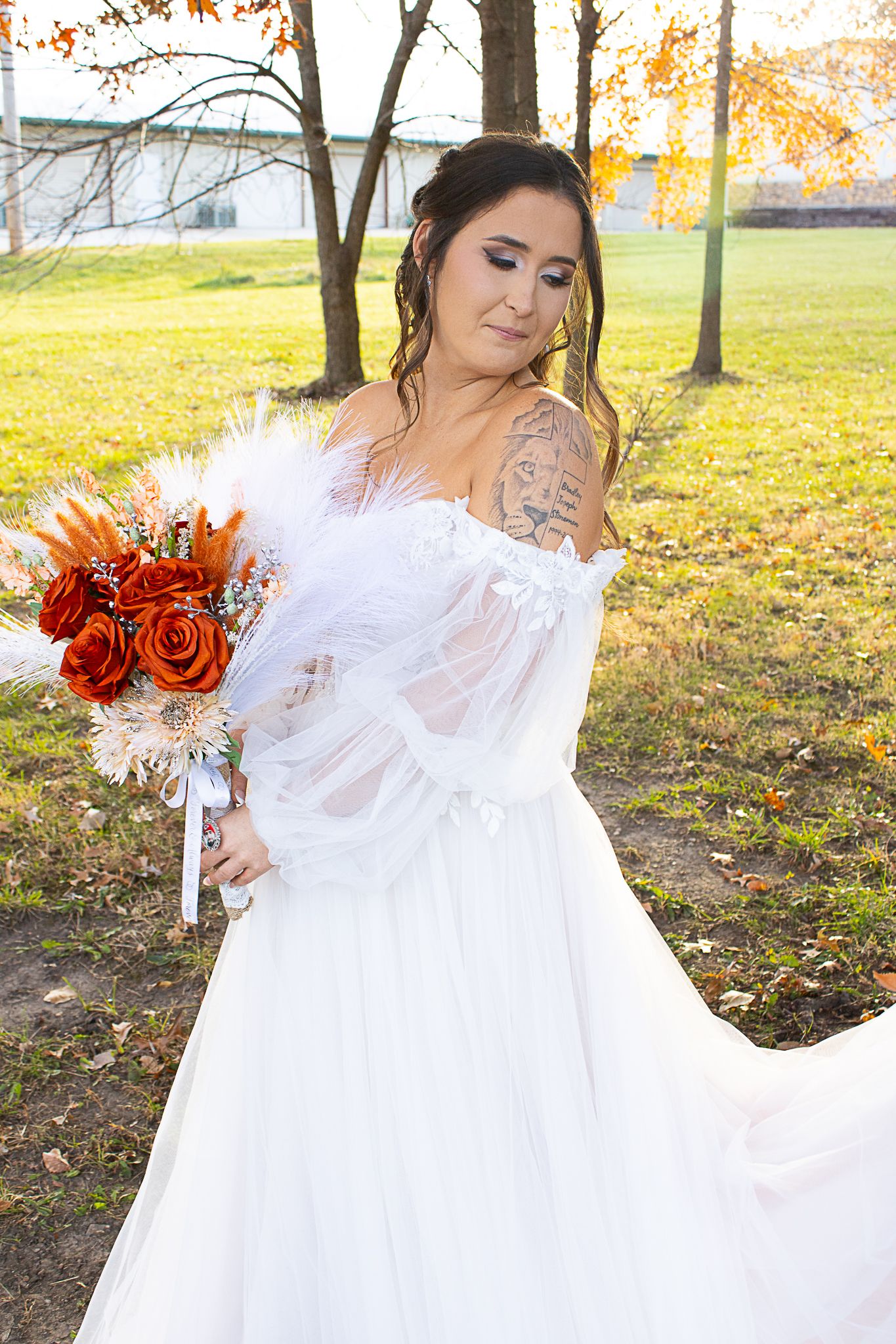 A bride in a white off-shoulder gown poses with her autumn bouquet featuring red roses.