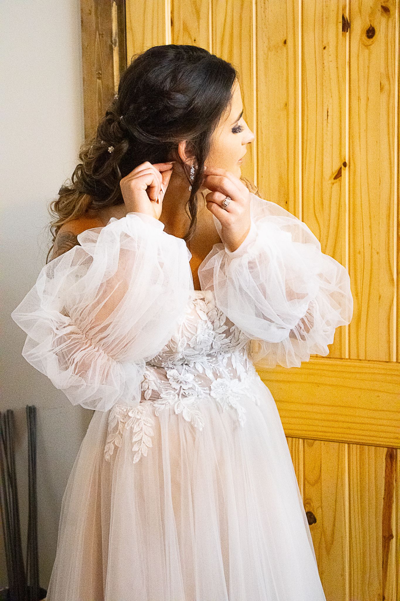 A bride in a white lace dress with puffy sleeves putting on earrings against wooden paneling.