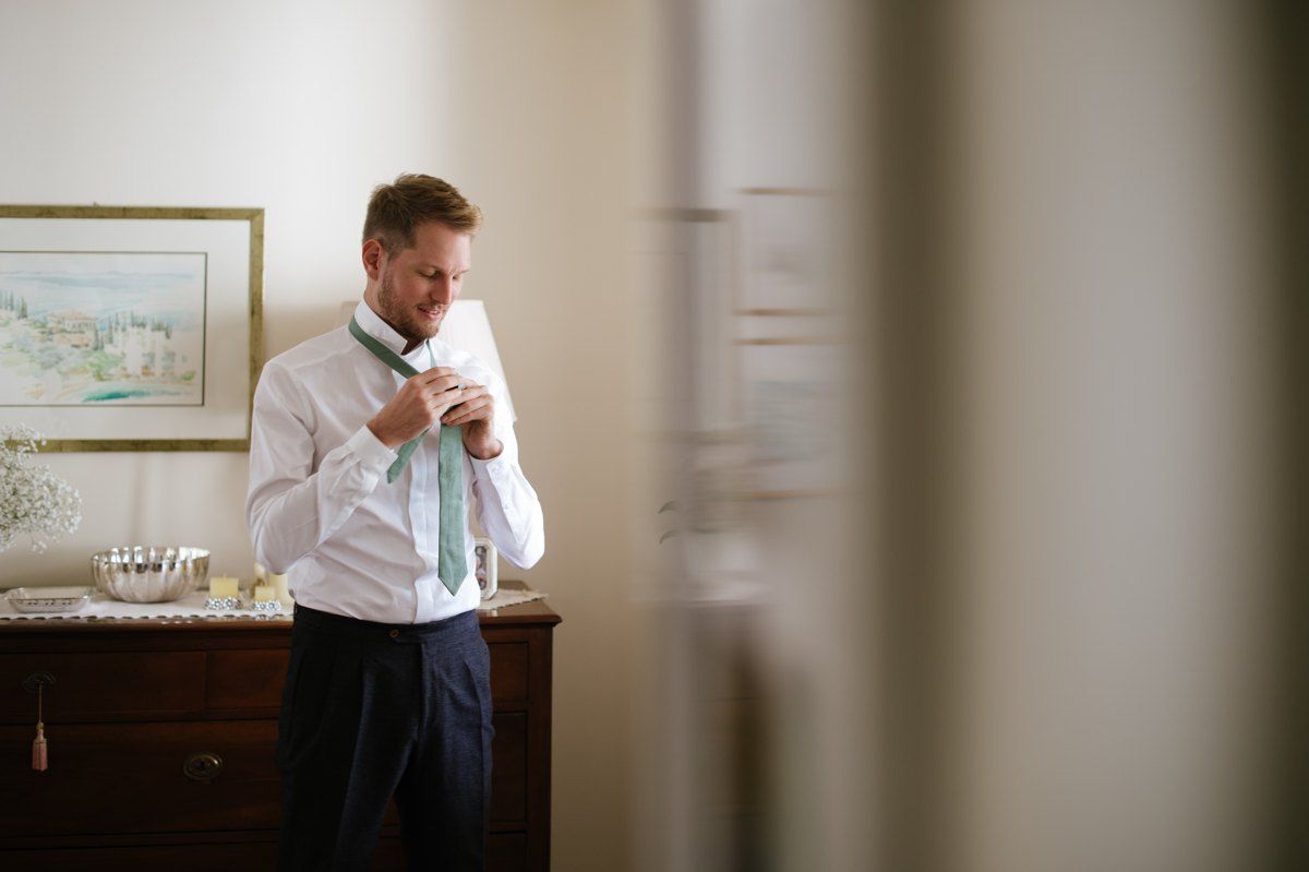 A person in white adjusts their tie while standing near a dresser.