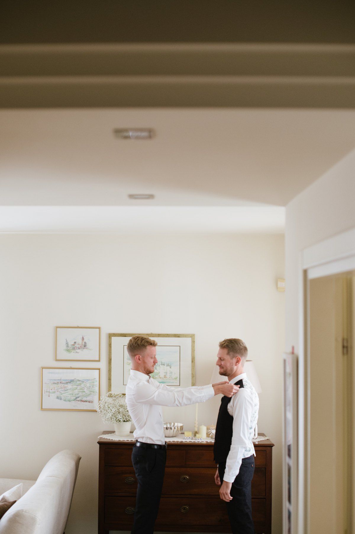 Two people assist with formal attire preparations near a dresser.