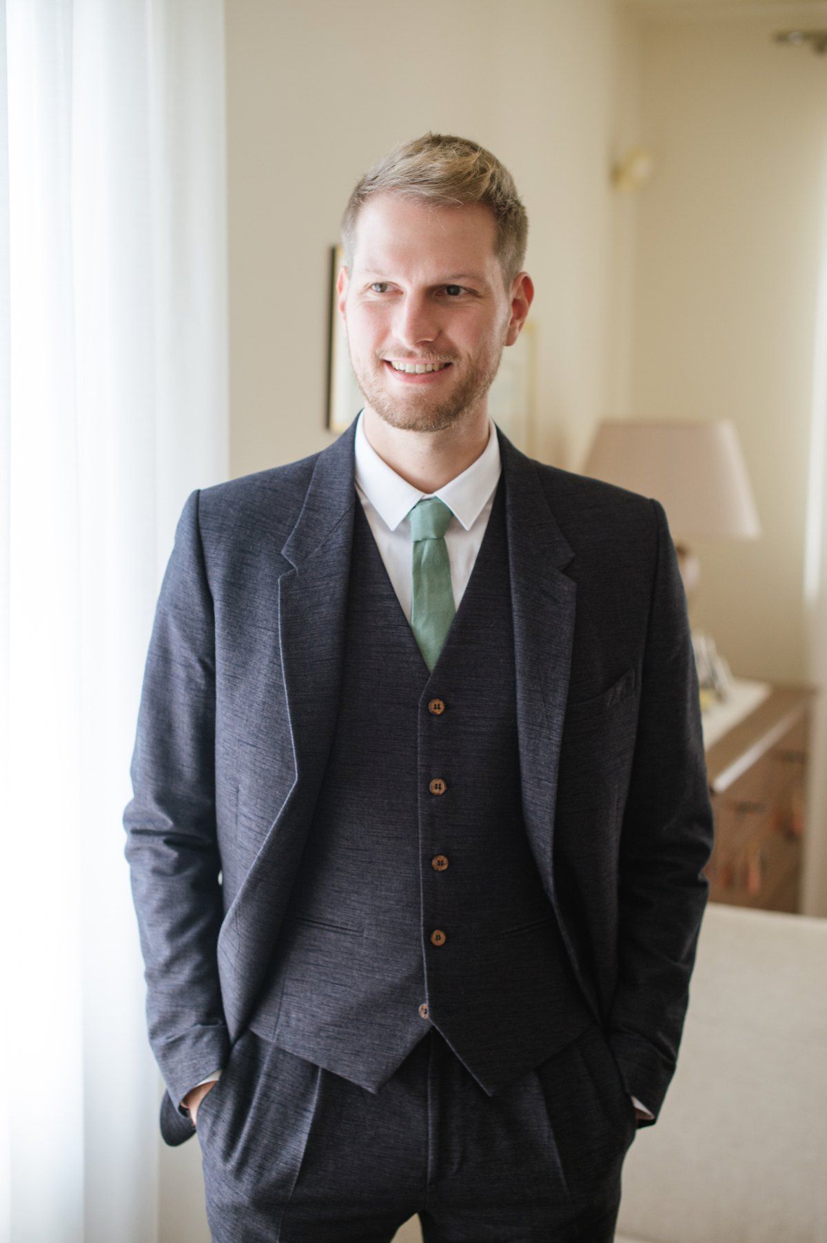 A person wearing a three-piece dark suit with mint green tie stands by a window.