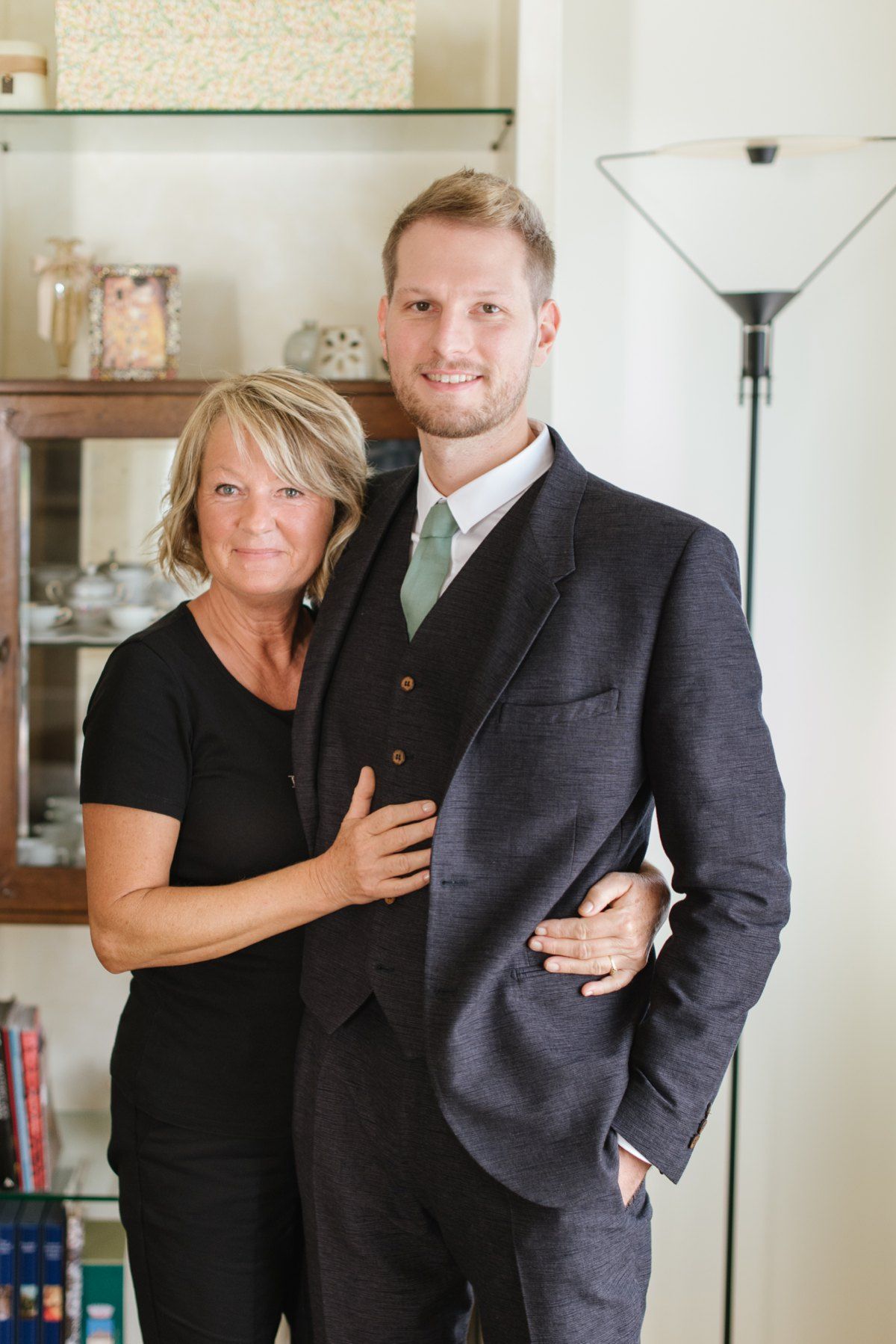 A pair posed together in home interior, person in dark suit with green tie beside person in black attire.