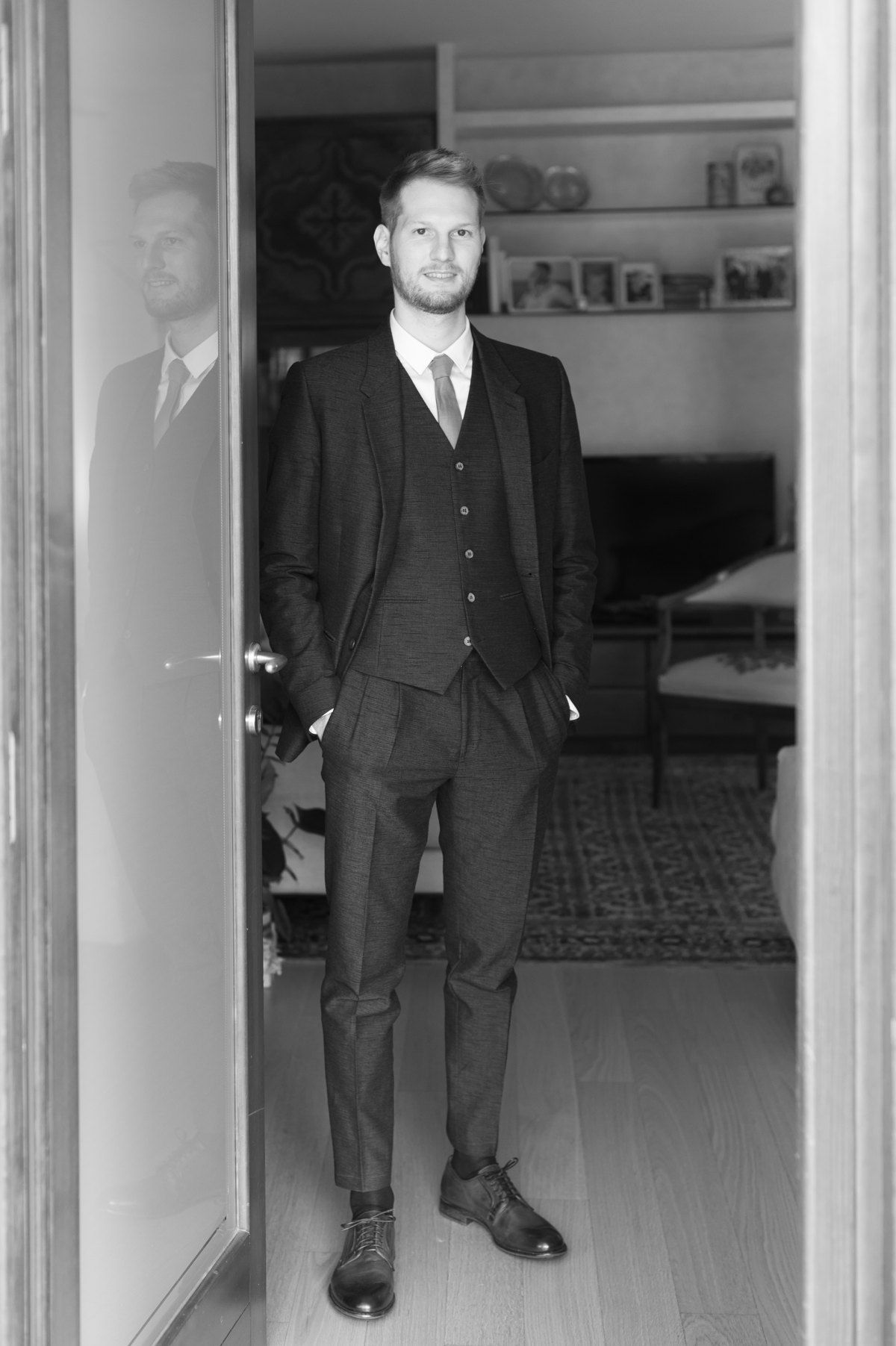 Black and white portrait of person in three-piece suit standing in doorway of modern home.