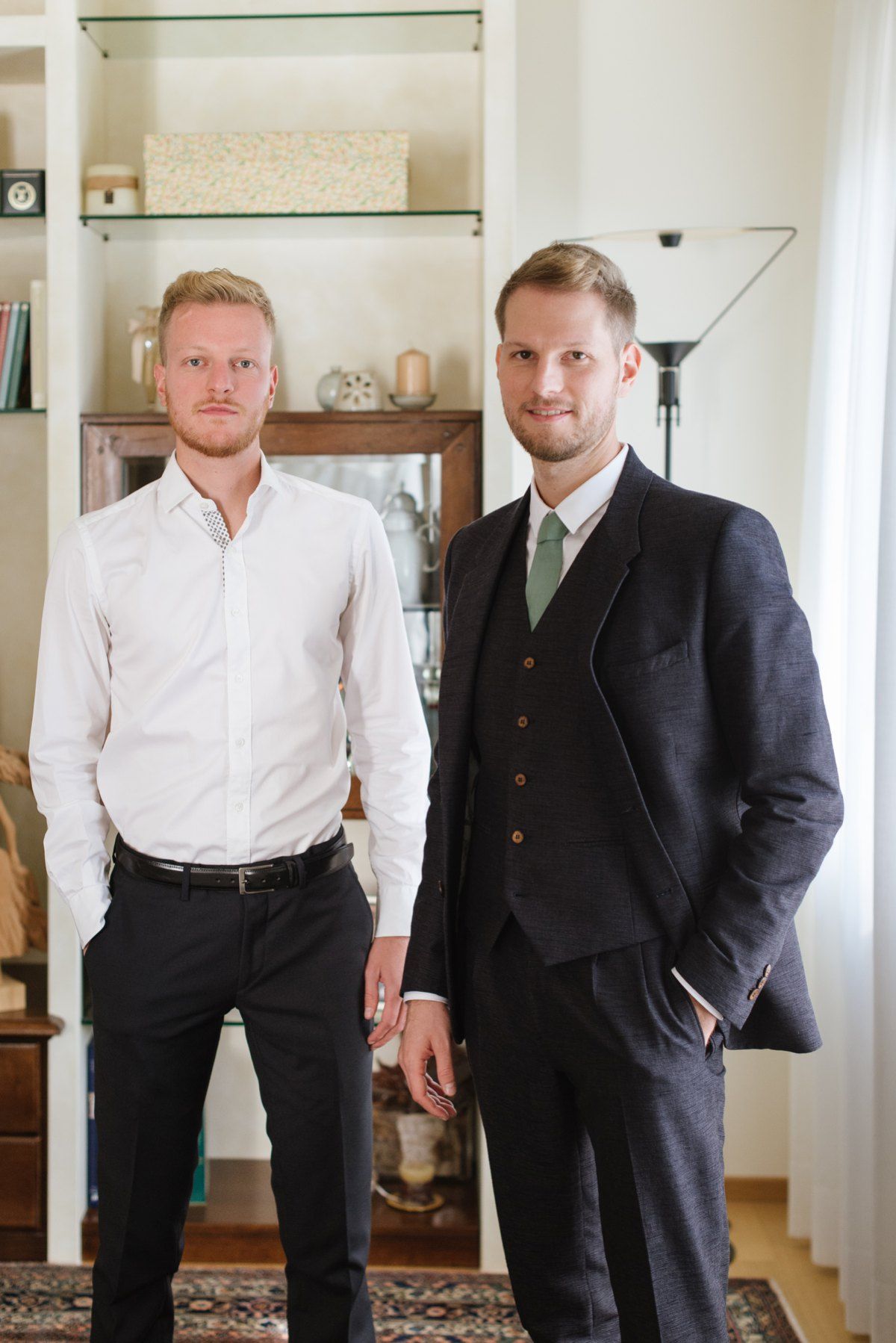 Two people posing in living room, one in white shirt and one in dark suit with green tie.