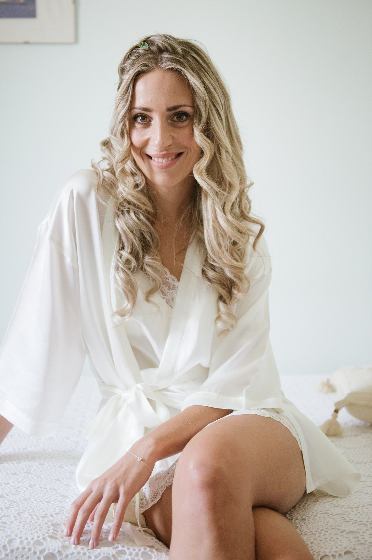 A bride wearing a white silk robe poses on a bed during wedding day preparations.