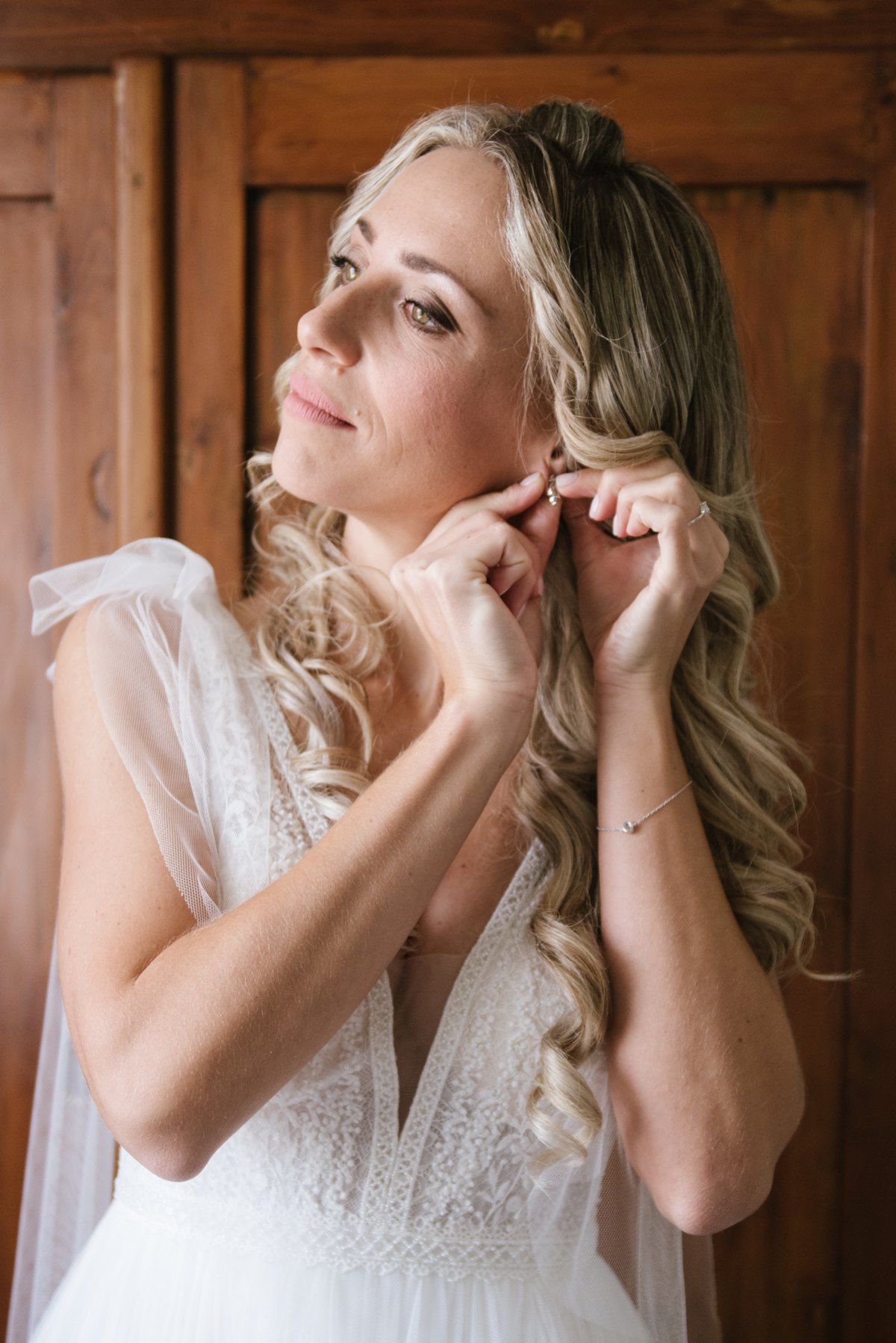 Bride in lace wedding dress adjusts her earring while getting ready.