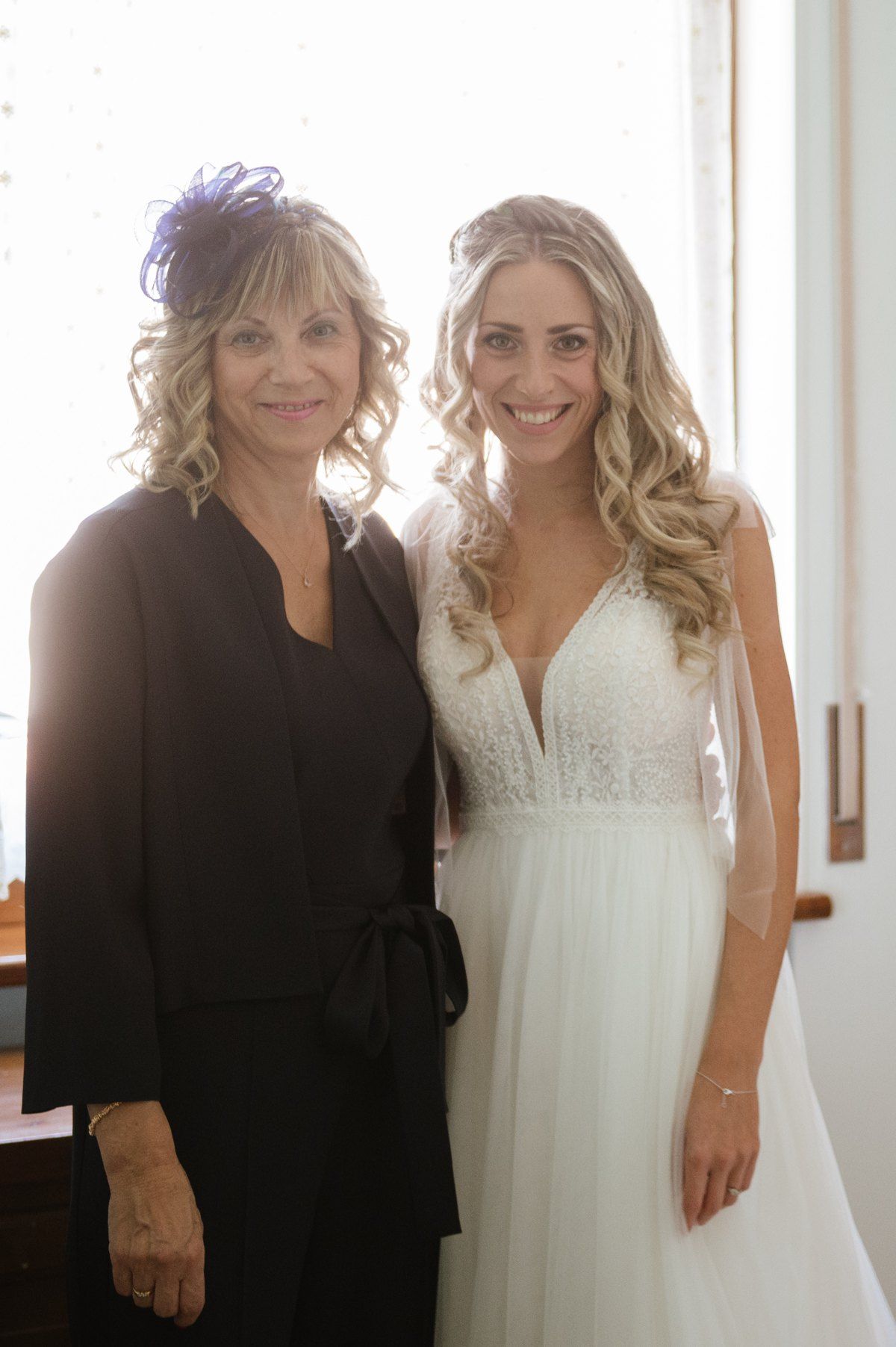 Two women stand together in a hallway, one in a black dress and one in a wedding gown.
