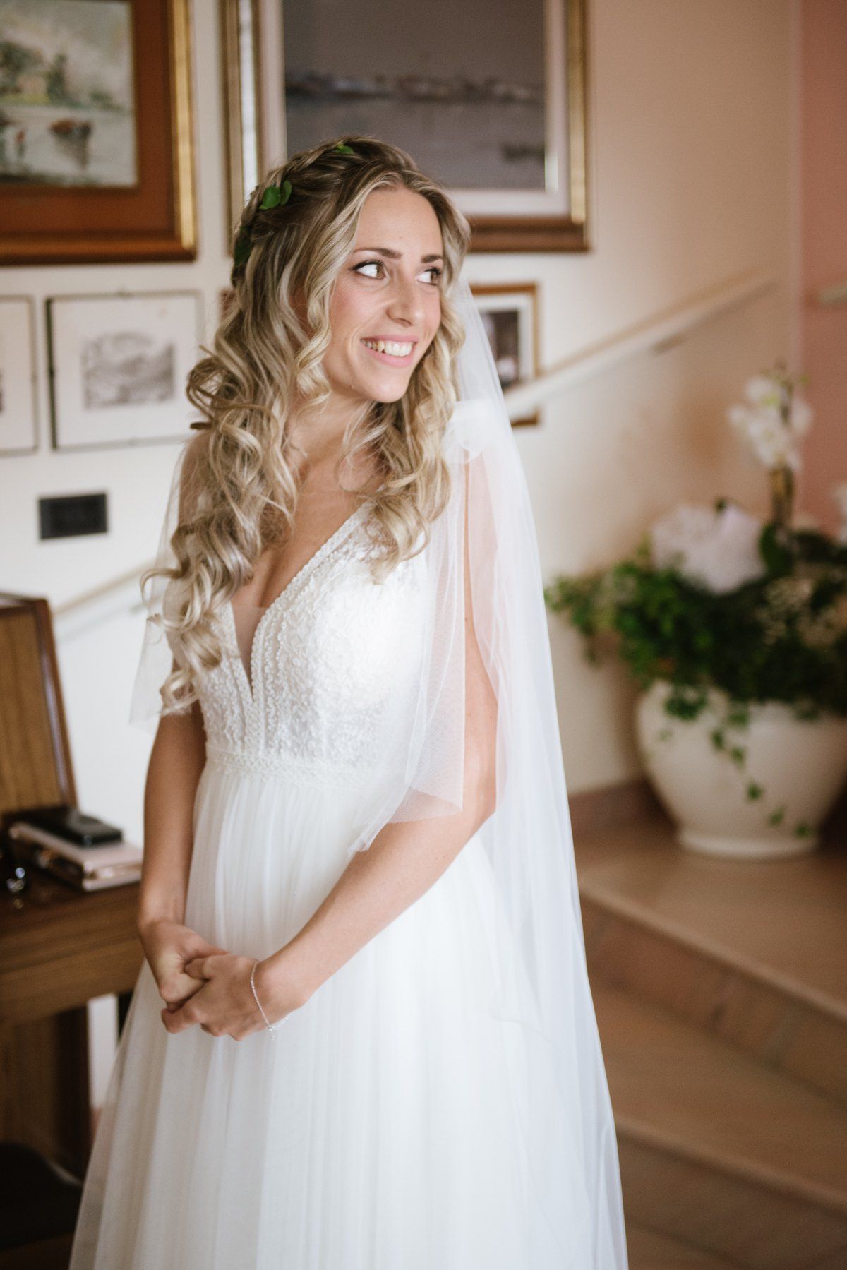Bride in flowing white gown with lace detail stands elegantly in home setting.