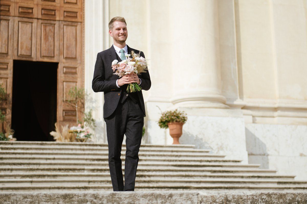 Wedding attendant in formal attire holding a floral bouquet on church steps.