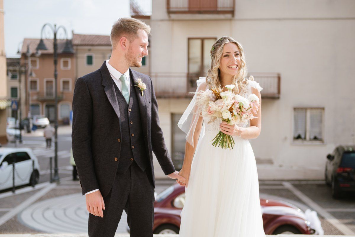 A couple stands together on their wedding day on an Italian street, with the bride holding a pale floral bouquet.