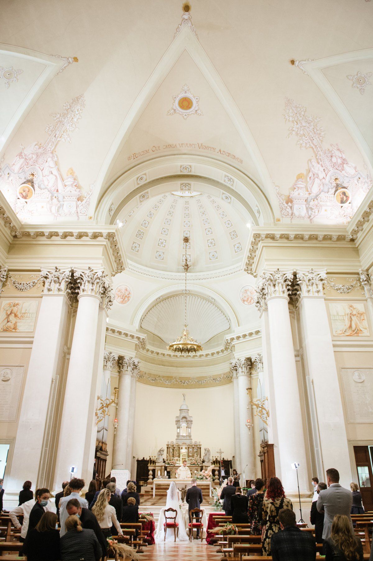 Grand white cathedral interior with ornate columns and vaulted ceilings during a wedding ceremony.
