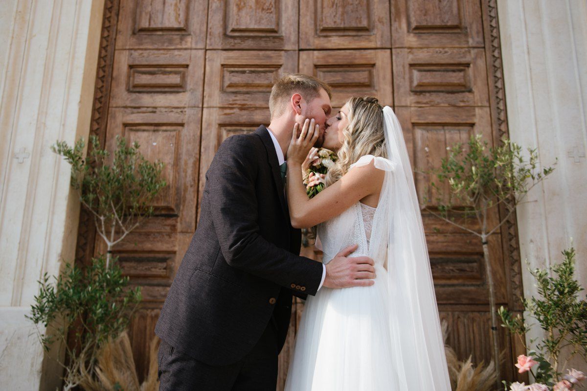 Newlyweds share their first kiss in front of ornate wooden church doors.