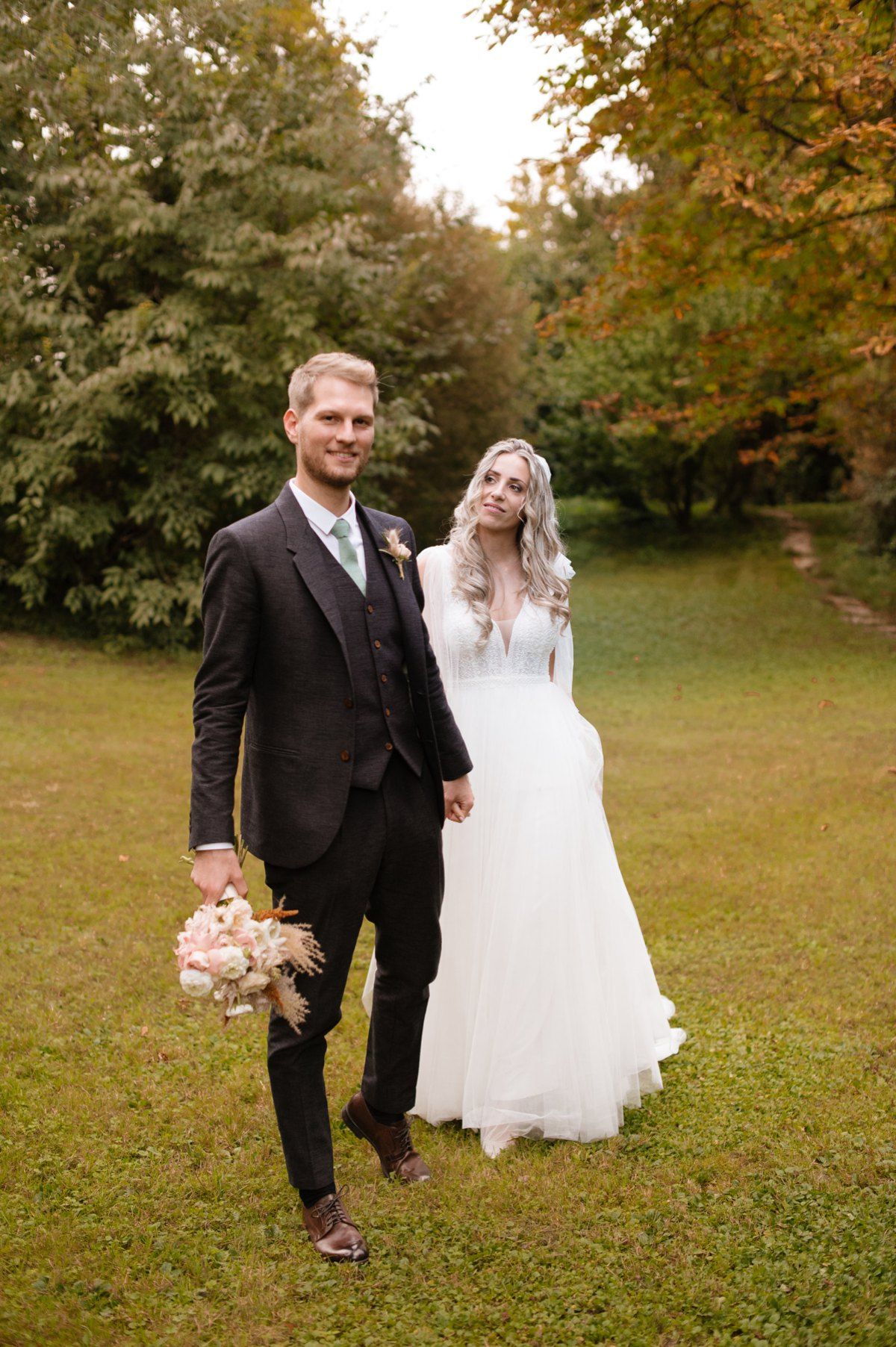 Wedding couple stands on grass holding bouquet with fall foliage in background.