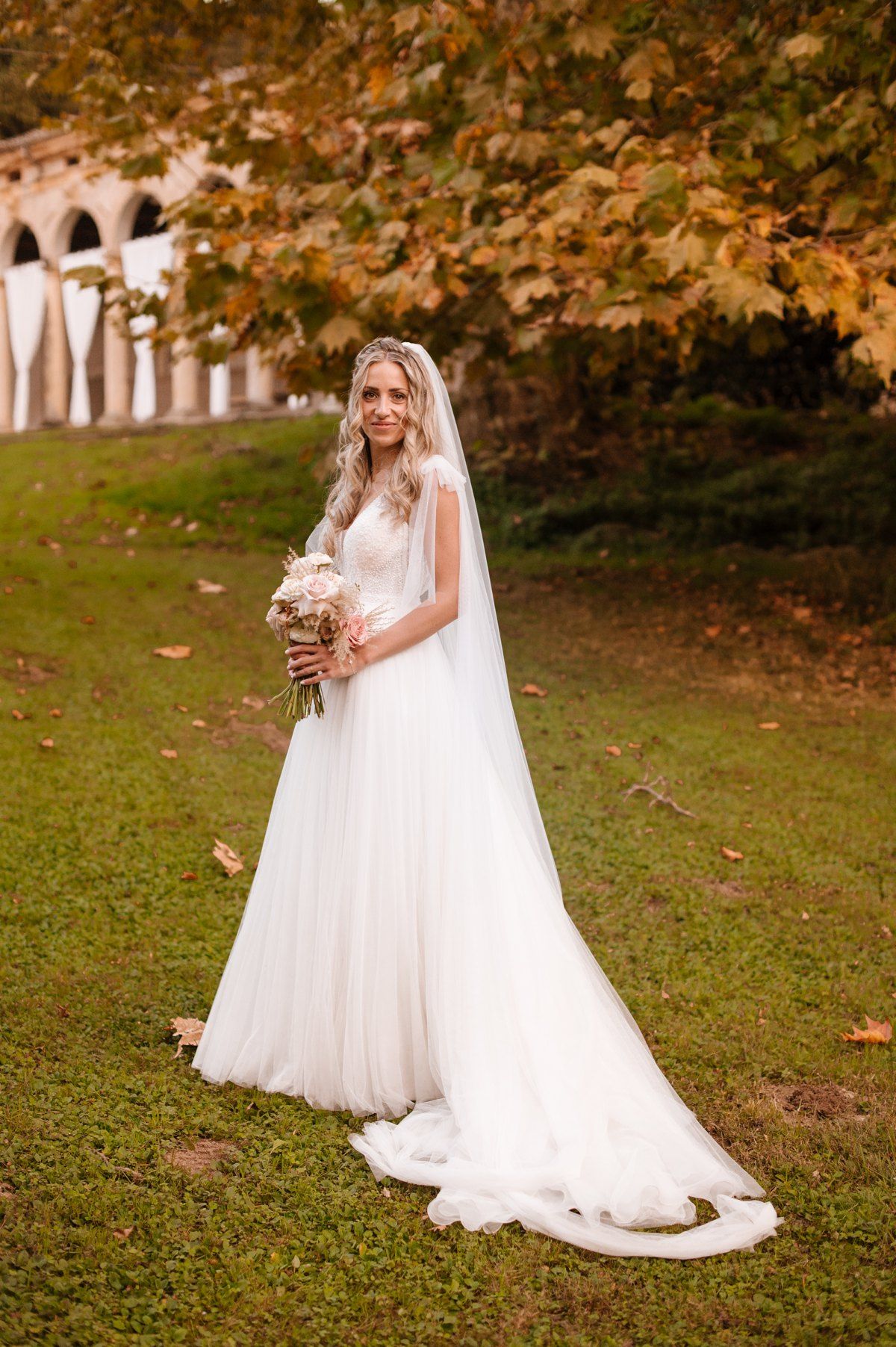 Bride in flowing white dress and veil poses with bouquet against autumn landscape with stone archway.