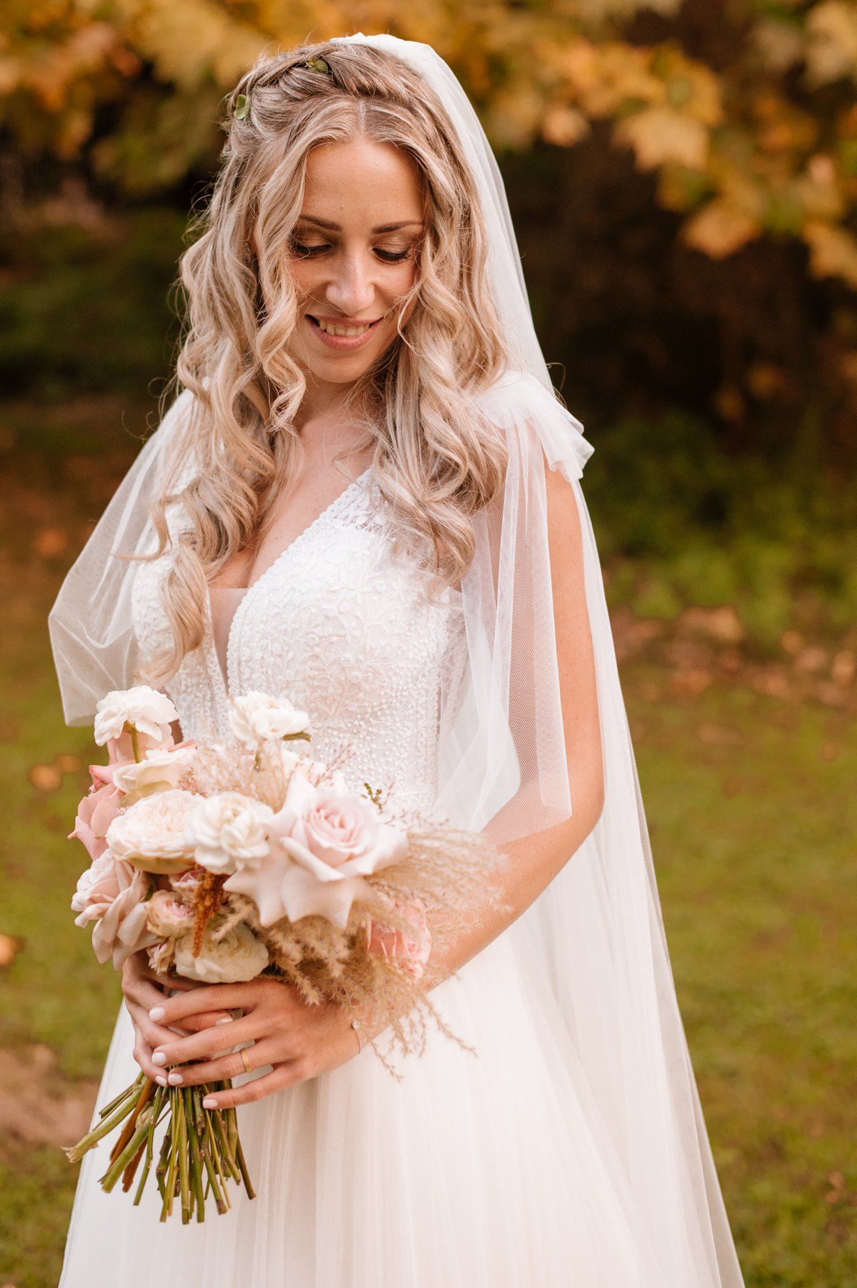 Bride in white gown holds blush pink bouquet while smiling with autumn colors in background.