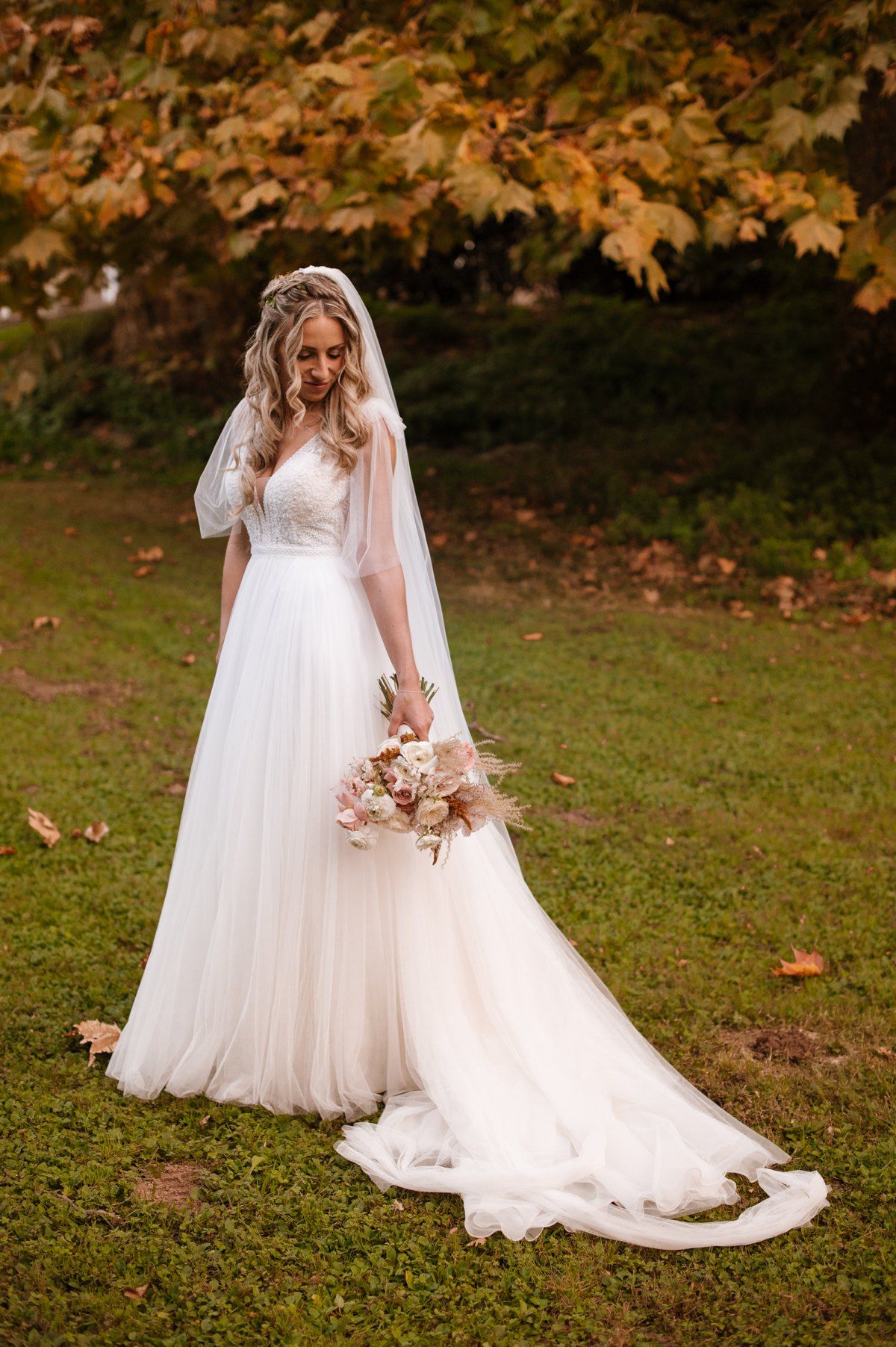 Elegant bride in flowing white gown and veil holds bouquet in autumn garden setting.