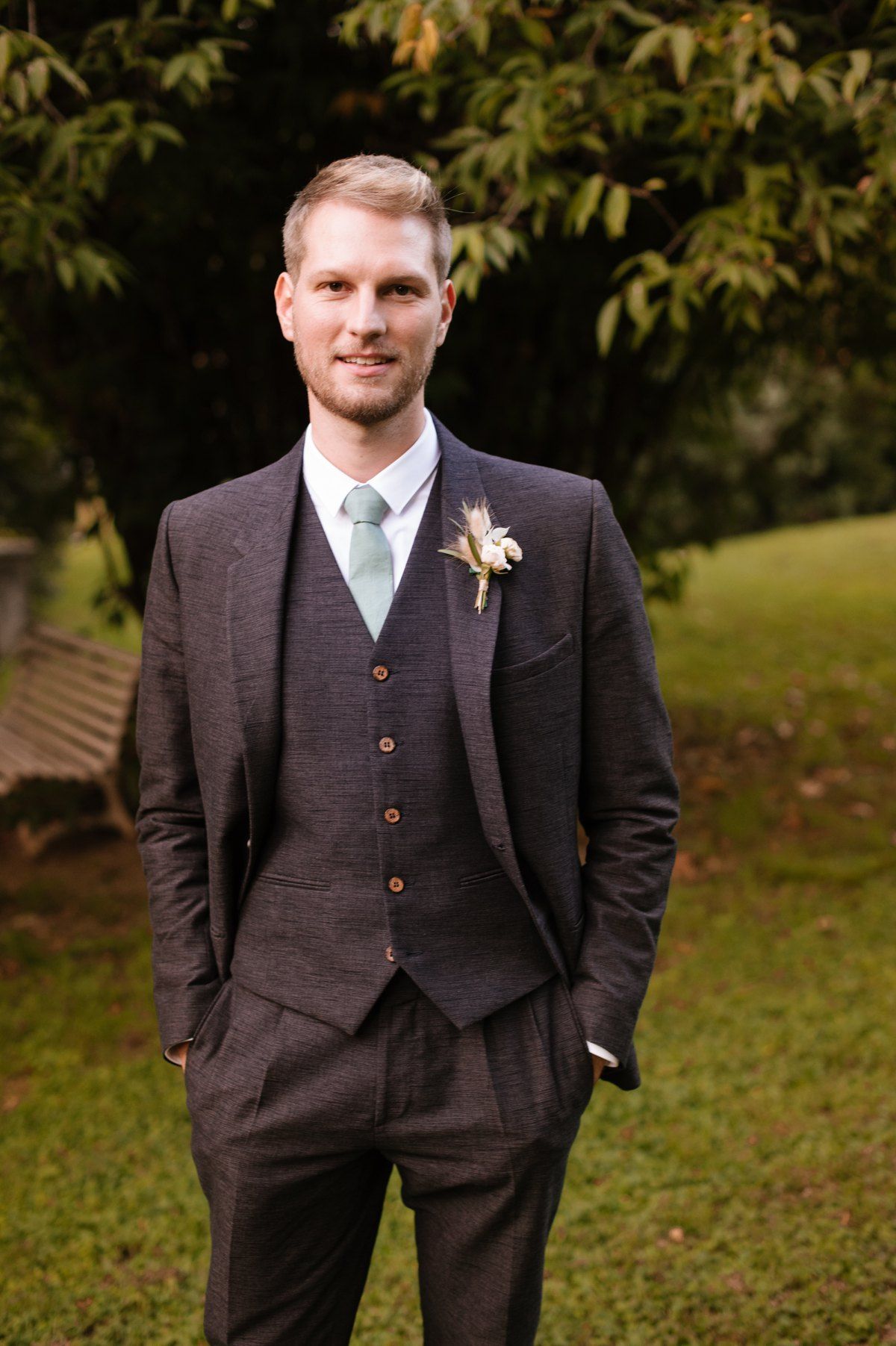 Groom wearing brown three-piece suit with light blue tie stands in garden setting.