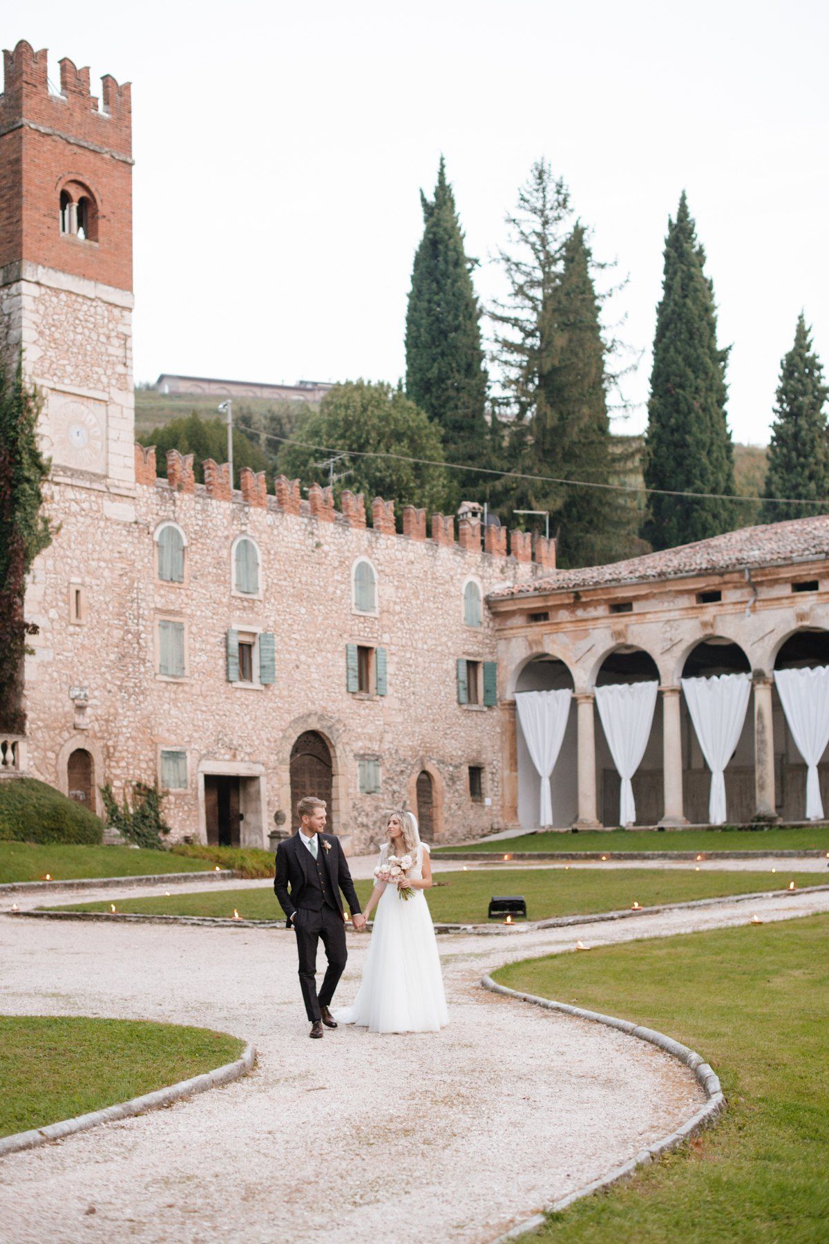 Couple walking along curved path at Italian castle venue with tall cypress trees and stone architecture.