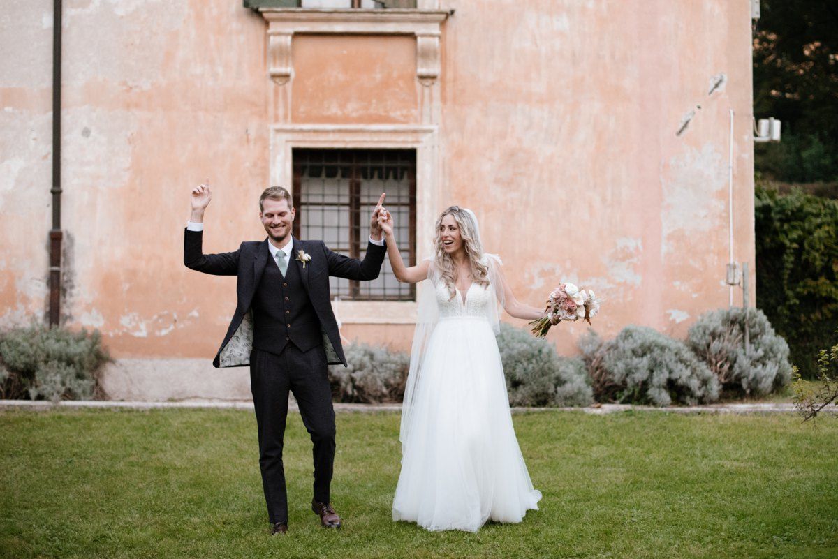Newlyweds celebrating joyfully on lawn in front of peach-colored villa.