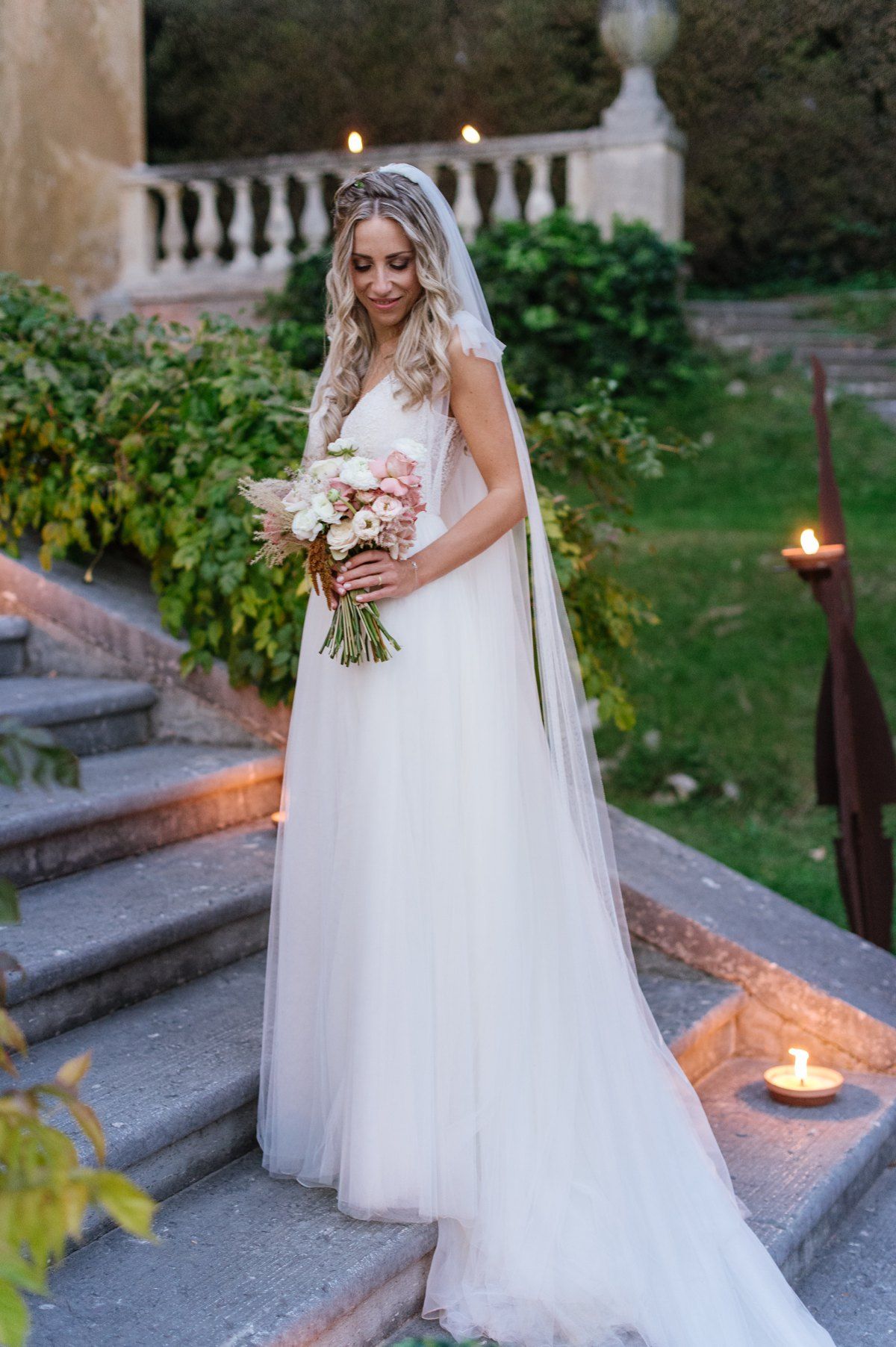 Bride in flowing white gown posing on candlelit stone steps with bouquet.