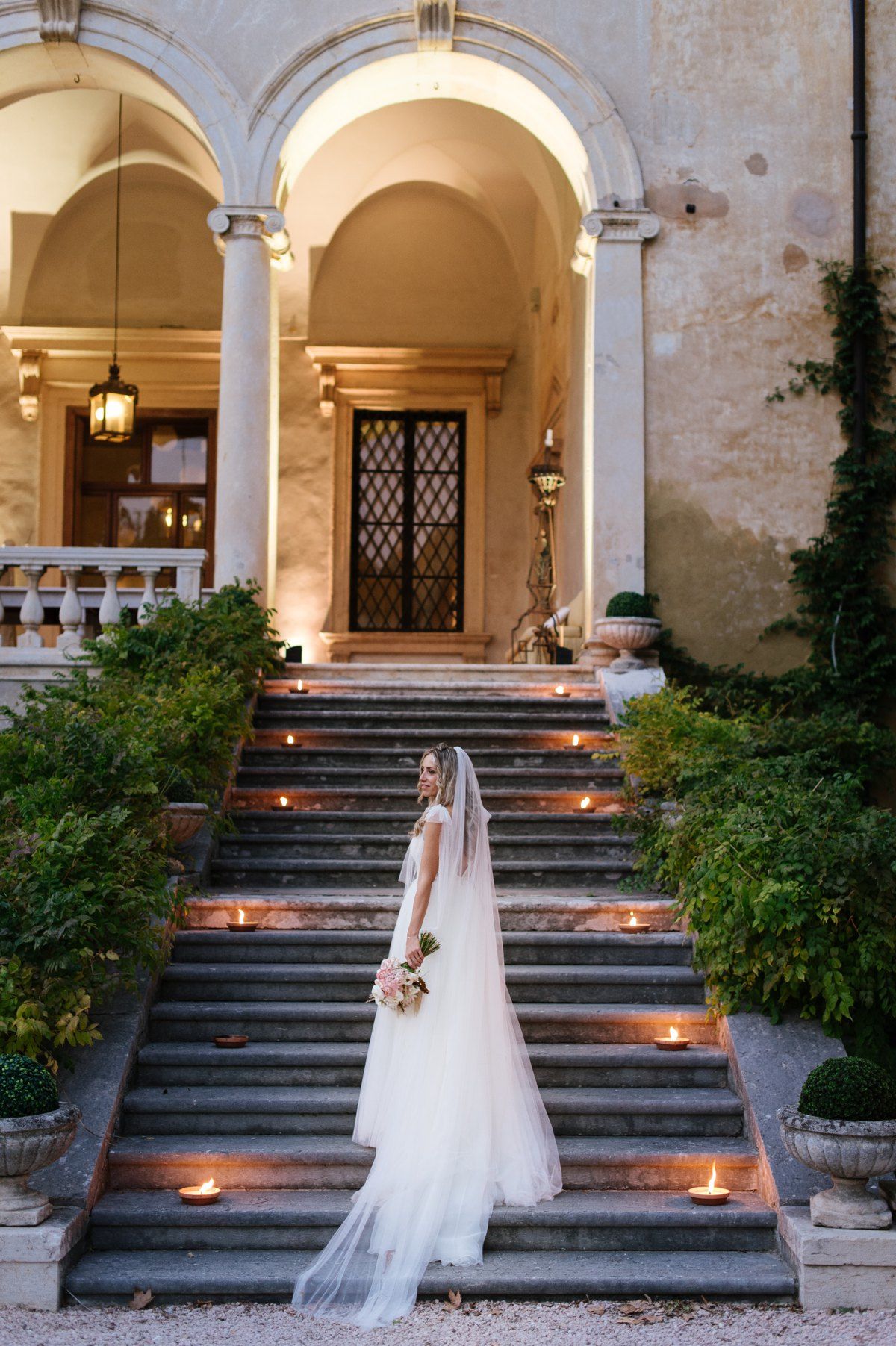 Bride standing on grand stone staircase lined with glowing candles and greenery.