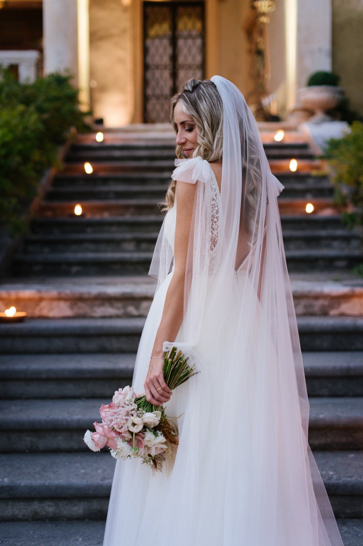 Bride in delicate lace gown with long veil holding pink bouquet on stone steps.