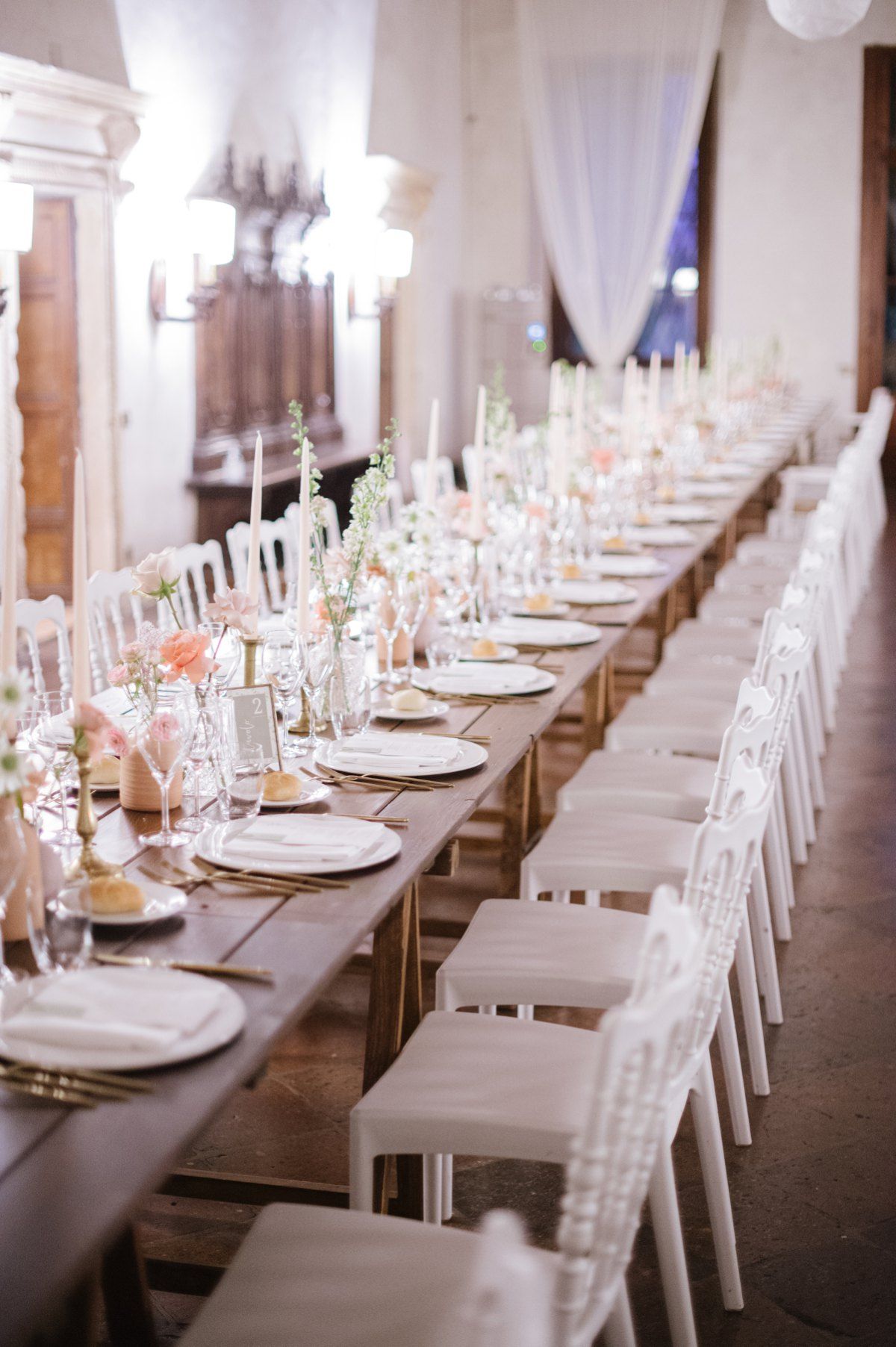 Long dining table set for wedding reception with white chairs and candlelit decor.