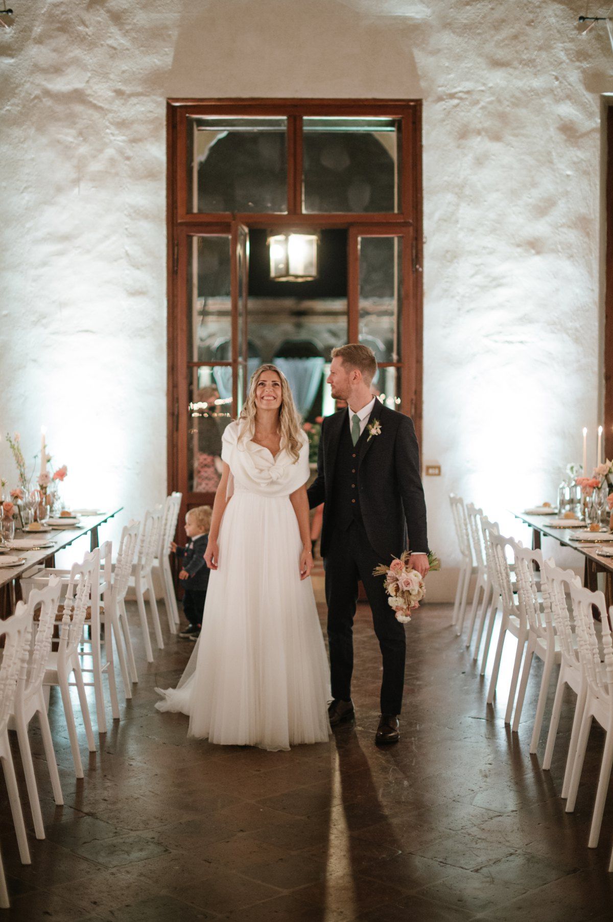 Wedding couple walking through elegantly decorated reception hall with white chairs.