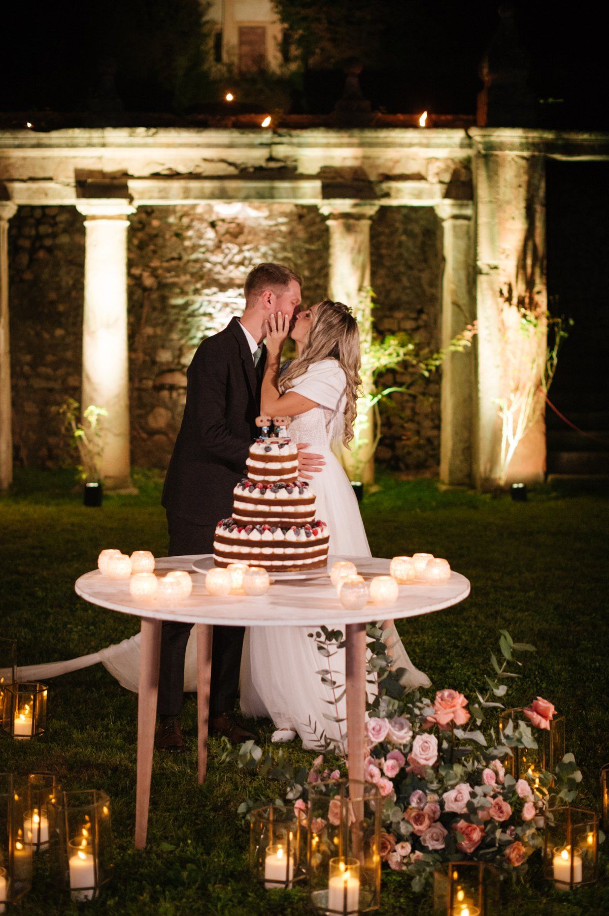 A romantic wedding cake scene with candles and stone columns illuminated at night.