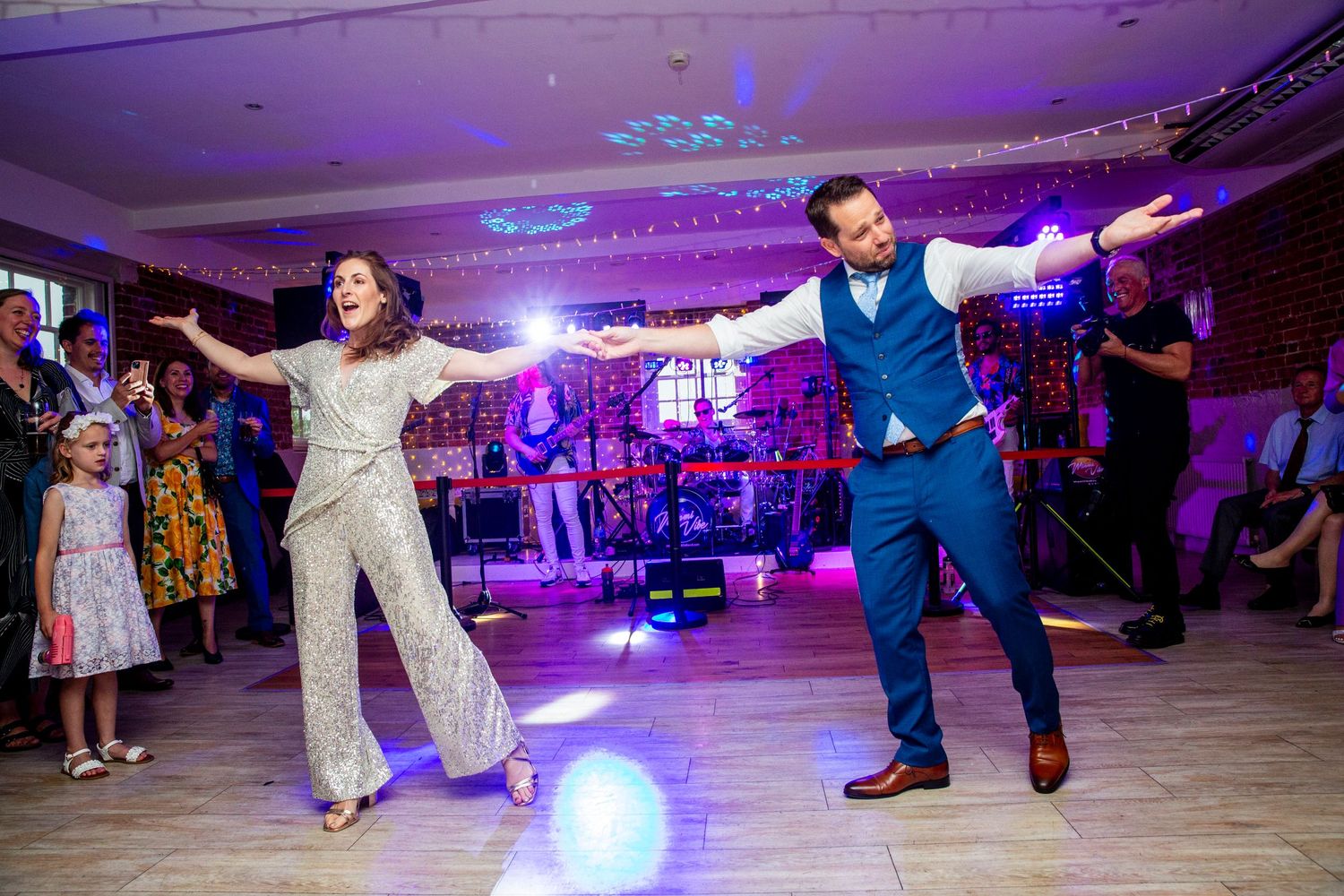 Wedding guests dance energetically on a purple-lit dance floor during a celebration.