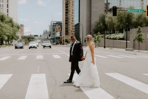 A couple in wedding attire crossing an urban crosswalk with tall buildings in the background.